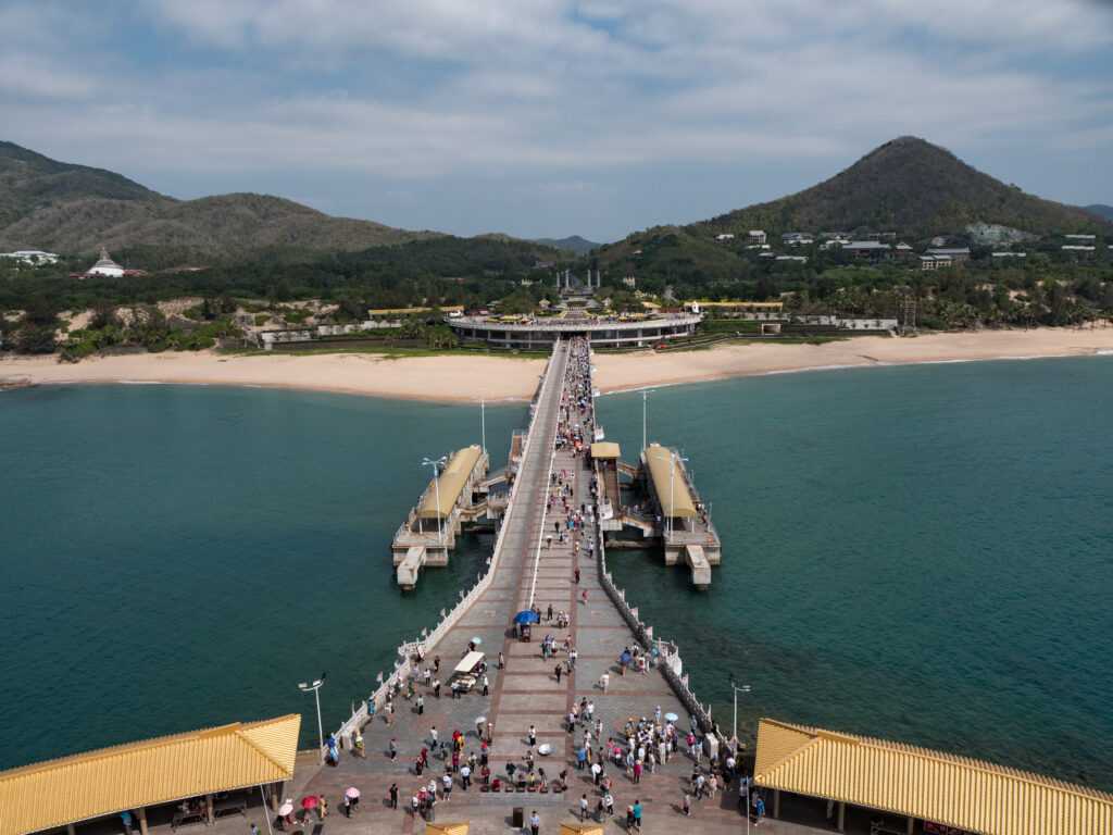 Crowded pier leading to a beachside complex with lush hills in the background, great to visit via car rental with driver.